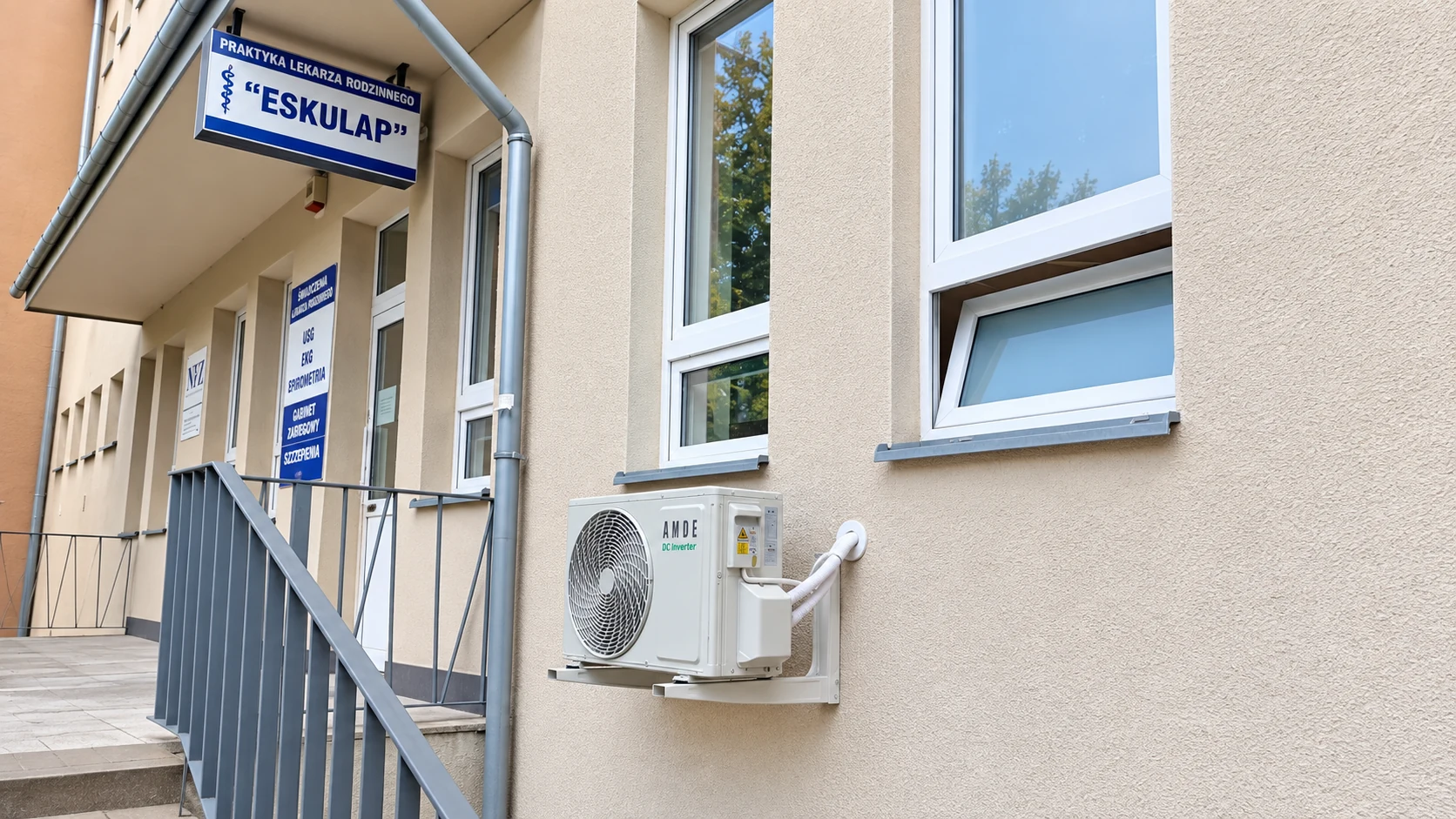Jednostka zewnętrzna klimatyzatora na elewacji pod oknem, obok schodów wejściowych.