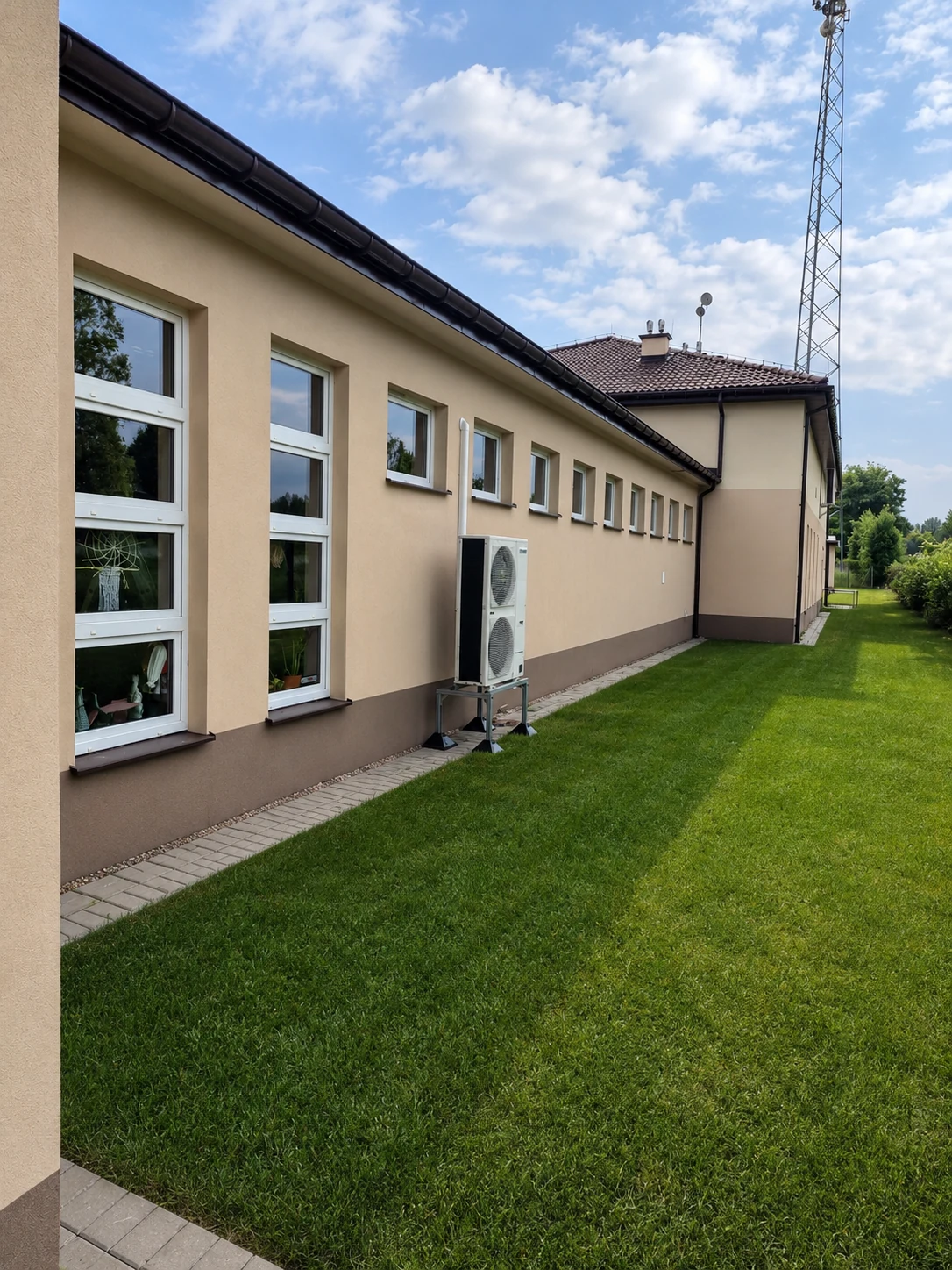 Dwuwentylatorowa jednostka zewnętrzna na stalowej podstawie obok jasnej elewacji.